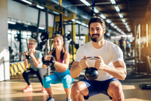 attractive-handsome-bearded-man-holding-kettlebell-doing-squats-fitness-group-with-two-girls-modern-gym